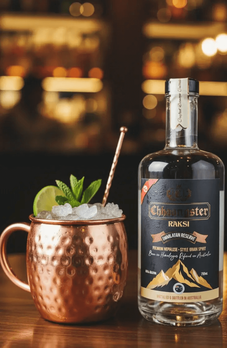 A Himalayan Mule cocktail in a copper mug with ice, lime, and mint garnish, served beside a bottle of Chhaangster Himalayan Reserve Raksi 700ml on a wooden bar counter.
